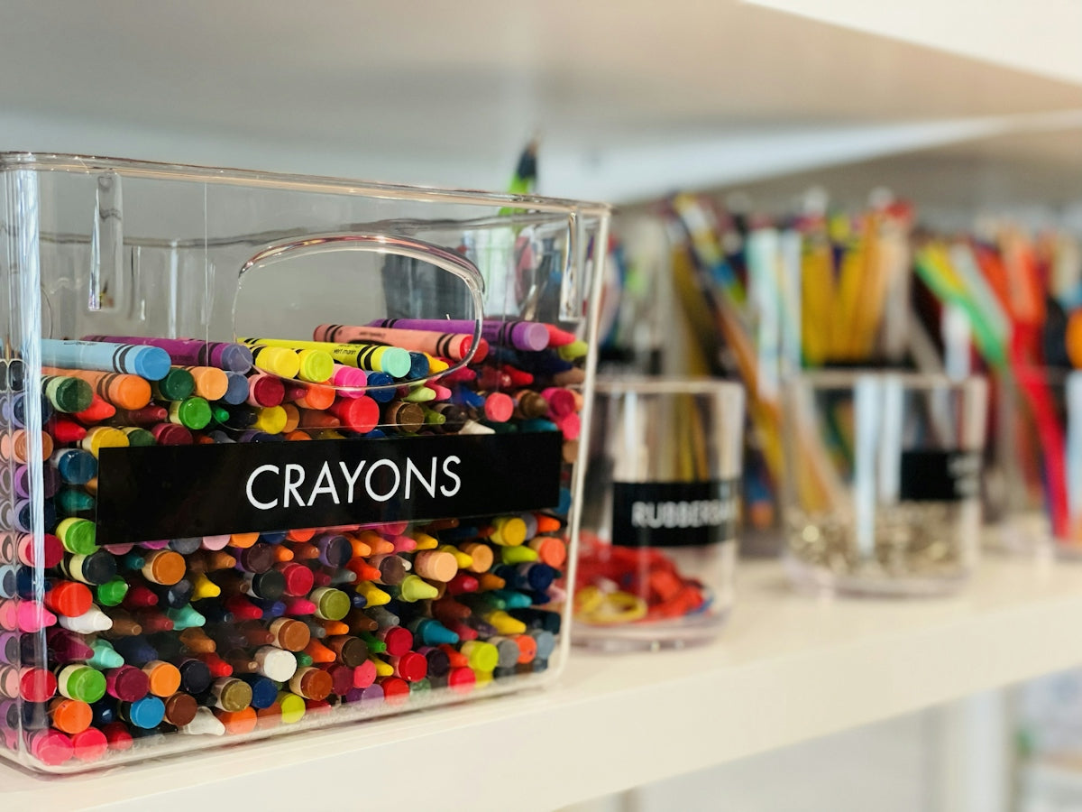 A clear plastic bin labeled CRAYONS filled with colorful wax crayons on a shelf